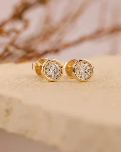 A pair of round bezel-set diamond stud earrings resting on pale stone