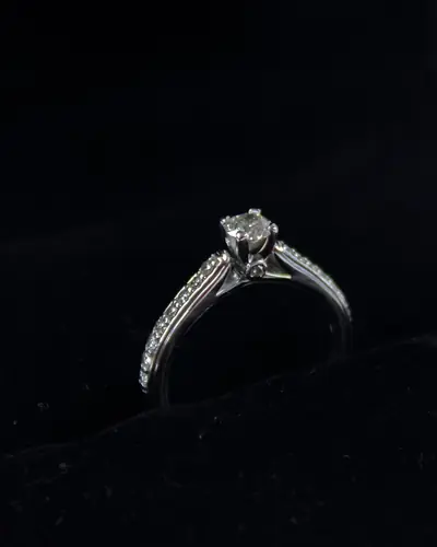 Solitaire engagement ring with a round diamond in six claws and diamond-set shoulders, on a black ground